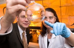 A male faculty member in a business suit is in a chemistry lab and holding a clear glass Florence flask containing a clear liquid. A female student wearing goggles, a white lab coat, and blue disposable gloves is pointing to the contents of the flask.