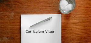 Image of a white piece of paper with "curriculum vitae" typed on it. The paper is on a wooden desk. A silver pen is lying on top of the paper. A glass of ice water is by the upper right corner of the paper.